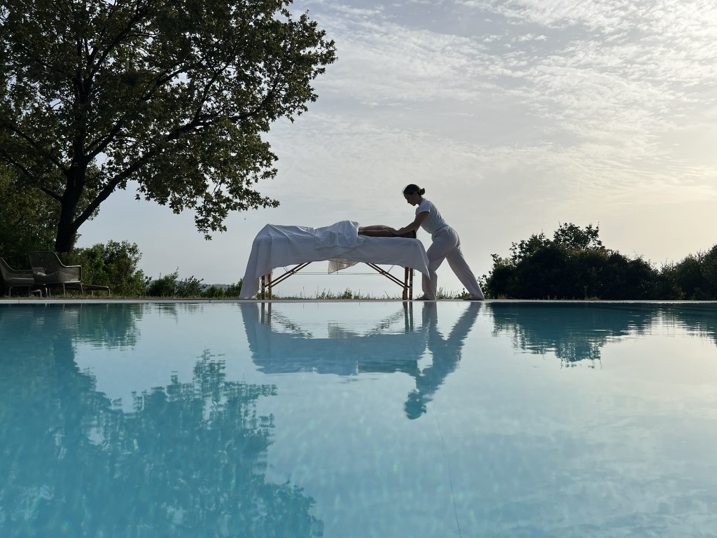A massage therapist performs a massage on a client outdoors by a pool, surrounded by lush greenery and a clear sky.