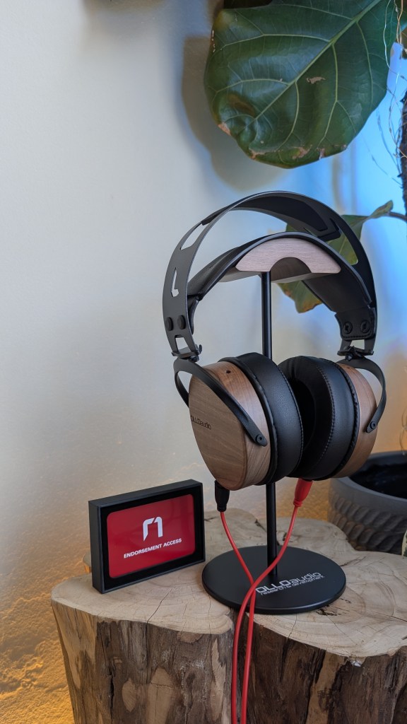 A pair of wooden and black over-ear headphones displayed on a stand next to a small red and black sign reading 'ENDORSEMENT ACCESS', placed on a wooden surface.