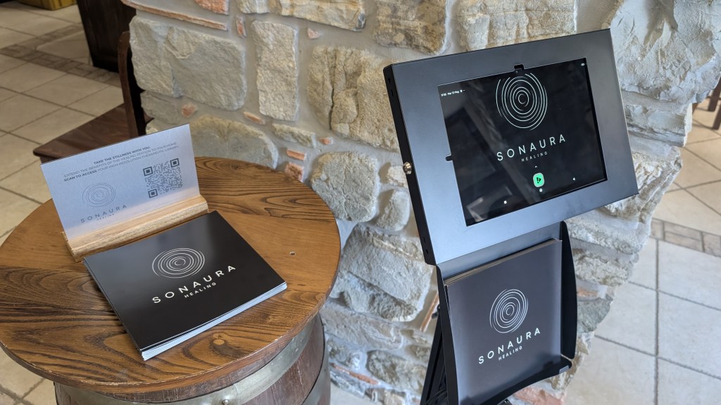 A wooden table displaying a black menu titled 'SONAURA HEALING' alongside a digital tablet with the same title, set against a stone wall.