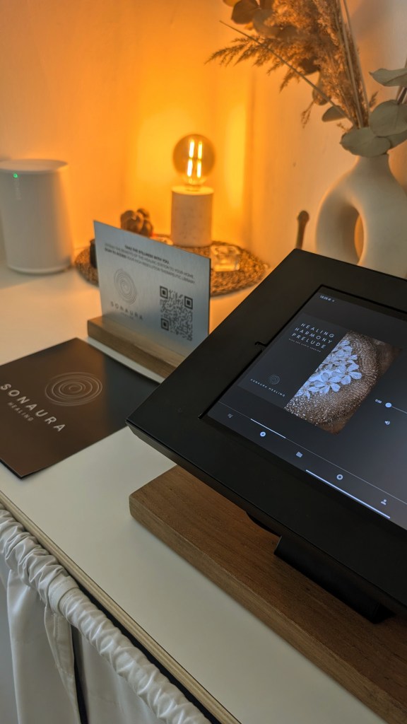 A modern digital display showing a serene image related to healing, positioned on a wooden stand, surrounded by soft ambient lighting and decorative elements, including a QR code and a menu.