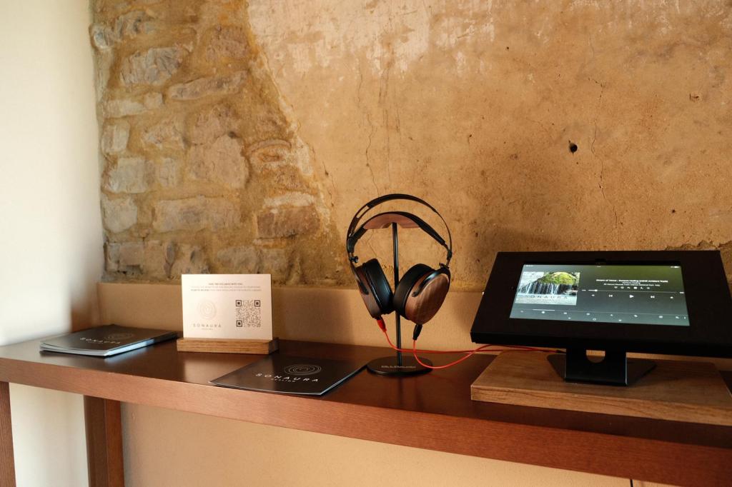 A wooden shelf displaying a pair of headphones on a stand, a touchscreen device showing content, and informational materials, against a stone wall.
